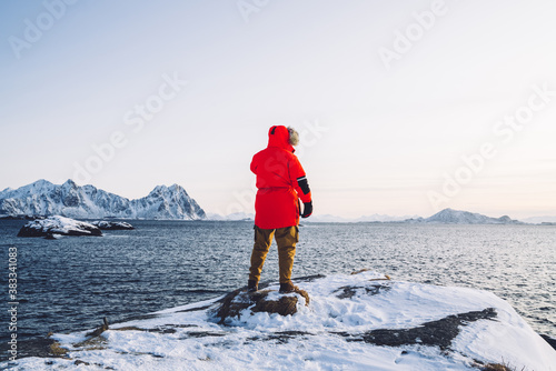 Back view of man in red winter coat standing near sea water in fiordland during vacation enjoying scenery, male fisher looking at calm water surface in lake on nordic environment