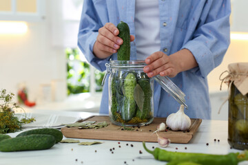 Wall Mural - Woman putting cucumber into pickling jar at table in kitchen, closeup