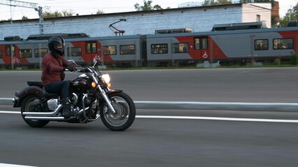 Motorcyclist in black helmet and red plaid shirt rides his burgundy chrome shiny motorcycle down the road with train on background. Same speed challange. Male biker rides chopper on the highway.