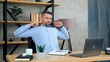 Tired business man SEO manager freelancer sits on chair at table in home office wearing wireless earphones doing warm up stretches body rubs tense muscles after long hard work at laptop computer