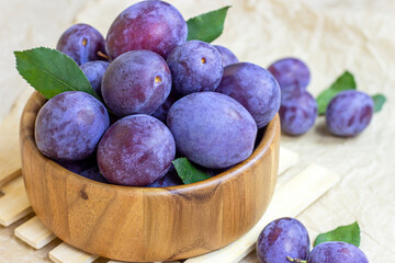 Fresh raw blue and violet plums in wooden bowl with green leaves on light paper background.