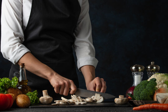 the professional chef in black apron cuts with knife mushrooms on black chopped board on dark blue b