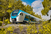 Modern And New Passenger Commuter Train In White And Blue Color Is Traveling On A Single Track Railway Line Between The Green Leaves On An Autumn Day.