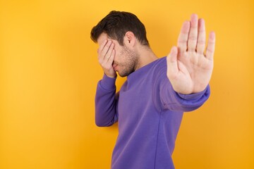 Wall Mural - Handsome man with sweatshirt over isolated yellow background covers eyes with palm and doing stop gesture, tries to hide from everybody.