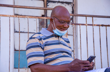 handsome elderly african man wearing face mask preventing, prevented himself from the outbreak in the society feeling excited about what he saw on his cellphone.