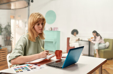 A creative young woman with blond hair sitting and working on her laptop while her colleagues sitting together on a background