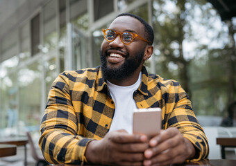 Young handsome African American man in stylish eyeglasses holding mobile phone, communication. Portrait of emotional guy using mobile application for online shopping, laughing, sitting in cafe