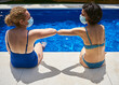© Cavan Images - Two women greet each other with the elbow. They are sitting on the edge of a pool