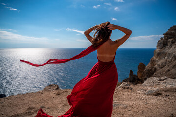From behind, a woman is seen in a red flying dress fluttering in the wind. In a straw hat, walking down the stairs against the background of the sea with rocks. The concept of travel