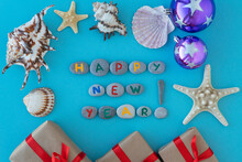 Flat Lay Of Inscription New Year Made With Colorful Letters On Gray Sea Stones Surrounded By Sea Shells And Stars, Gift Boxes Tired With Red Ribbons. Greeting Travelling Concept.