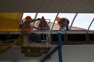 two workers repair the roof near the railway station. One worker is in a lift with a cradle, the second is on the roof, with insurance and a grinder tool