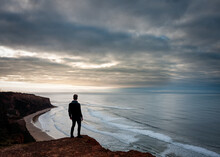 Hiker Standing On A Great Ocean Road Cliff Overlooking A Moody Beach