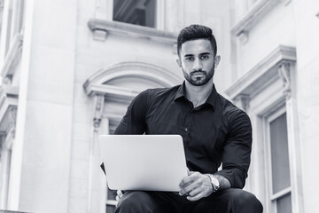 Young East Indian American Man with beard, studying, working in New York City, wearing black shirt, holding laptop computer, sitting outside old style office building, looking up. Black and White..