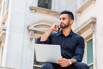 Young East Indian American Businessman with beard working in New York City, wearing black shirt, holding laptop computer, sitting outside old style office building on campus, talking on cell phone..