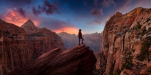 Adventurous Woman At The Edge Of A Cliff Is Looking At A Beautiful Landscape View In The Canyon During A Vibrant Sunset. Taken In Zion National Park, Utah, United States. Sky Composite Panorama