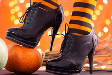Witch Legs In Striped Stockings And High Heel Shoes With Pumpkins On An Orange Background, Bokeh. Halloween. Copy Space.