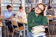© JackF - Annoyed angry charming girl sitting with pile of books in high school library on background of working fellow students