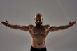 Professional bodybuilder posing. Muscular strong bald man with naked torso standing with outstretched arms isolated over grey background