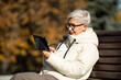 an adult woman in age sits on a bench with a tablet in her hands in a park in autumn