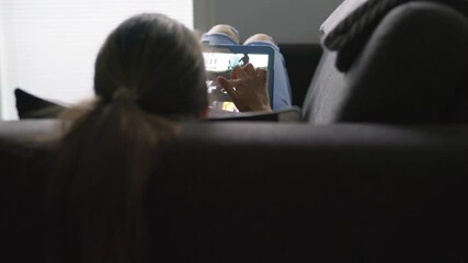 Wall Mural - Back view of woman using tablet and lying on couch in home living room. Person using movie stream service online and choosing series or film to watch. Streaming on demand video or episode of a tv show