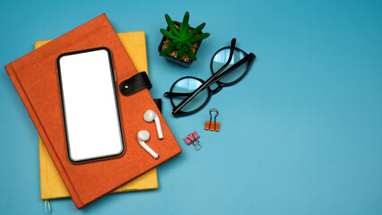Wall Mural - Flat lay, Workspace back to school and education concept on blue table desk with blank screen phone on stack of book, earphone and eyeglass, green plant, Top view with copy space