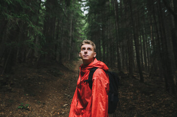 Portrait of a handsome pensive caucasian young man looking up, feeling curious and adventurous, wearing a red waterproof coat and black backpack. Mountain nature, fir forest.