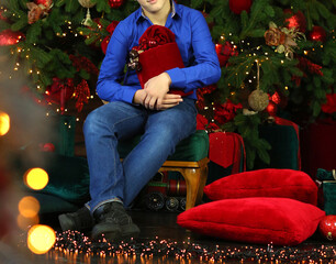 Caucasian teenage boy in a blue shirt sitting with a huge red box with gifts in a New Year's interior. New Year's and Christmas. Gifts concept.