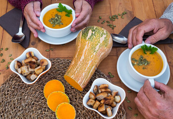 Close-up of two pairs of people hands ready to eat a pumpkin cream for an healthy and vegan or vegetarian lifestyle. Rustic and recycled wood table with pumpkin seeds and  bread croutons