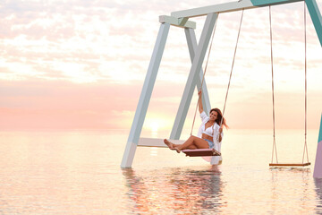 Wall Mural - Young woman enjoying sunrise on swing over water