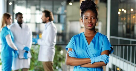 Portrait of African American happy beautiful woman physician in gloves smiling to camera in clinic. Joyful pretty female doctor or nurse in hospital. Mixed-races docs talking on background.