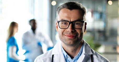 Wall Mural - Portrait of Caucasian man physician in white gown and glasses looking at camera, smiling and standing in clinic. Handsome male doctor in hospital. Doctors on background. Medicine concept.