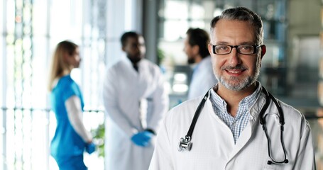 Wall Mural - Portrait shot of Caucasian senior man physician in white gown, stethoscope and glasses looking at camera and smiling in clinic. Handsome male doctor in hospital. Doctors on background Medicine concept