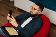 © eduard - Handsome man in blue shirt and glasses sitting armchair and holding a mobile phone in hands, laptop and notebook on the knees.