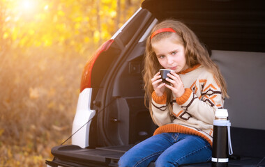 Wall Mural - Cute little girl drinking hot tea in car trunk on autumn background