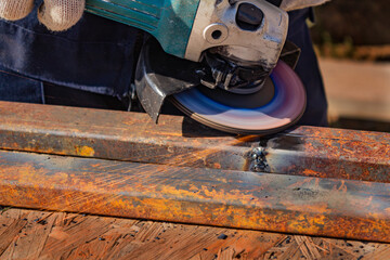 A worker in overalls is grinding a weld seam on a steel square profile pipe with an angle grinder equipped with a flap disc