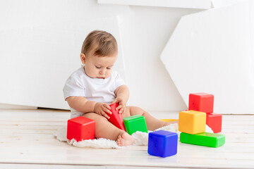 cute little baby six months old in a white t shirt and diapers playing at home on a Mat in a bright room with bright colored cubes