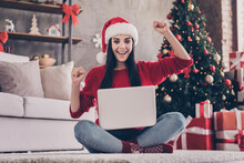 Photo Of Charming Excited Girl Sit Carpet Hold Laptop Raise Fists Wear Santa Cap Red Pullover Jeans Socks In Decorated Living Room Indoors