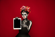 Holding Tablet. Young Girl Like Santa Muerte Saint Death Or Sugar Skull With Bright Make-up. Portrait Isolated On Red Studio Background. Celebrating Halloween Or Day Of The Dead. Copyspace On Screen.