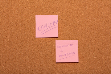 Covid-19 and Prevention is Protection handwritten on two pink stickers on a cork notice-board.