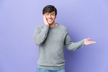 Young cool man holds copy space on a palm, keep hand over cheek. Amazed and delighted.