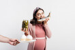 © LIGHTFIELD STUDIOS - pregnant woman holding jar with pickled cucumbers and eating dried fish while looking at cupcakes on plate in male hand isolated on white