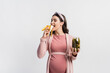 © LIGHTFIELD STUDIOS - joyful pregnant woman eating banana and holding jar with canned cucumbers isolated on white