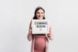 © LIGHTFIELD STUDIOS - joyful pregnant woman holding board with coming soon lettering isolated on white