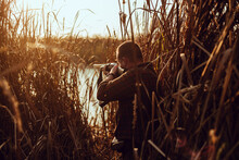Young Hunter Man With A Shotgun Hiding In The Reeds Near The Pond, At The Duck Hunt - Photo With Selective Focus