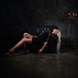 Sexy fit blod model with long legs in high heels relaxing and lying in fashion armchair in black dress and posing on dark dramatic background in dark shadow light. Mystic atmosphere. Stunning