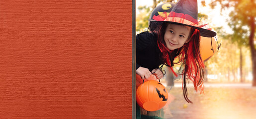 Wall Mural - Halloween trick or treat.Happy girl wearing a witch's hat, with a pumpkin handbag, peeps out from behind a Orange wall. Funny kid in carnival costumes outdoors.