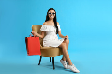 Wall Mural - Beautiful young woman with paper shopping bags in armchair on light blue background