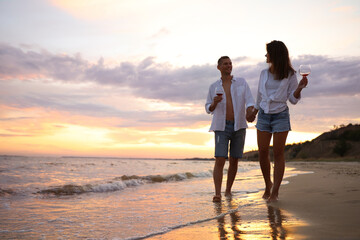 Canvas Print - Lovely couple with glasses of wine walking on beach at sunset. Space for text