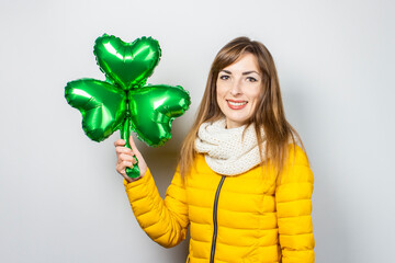 Woman with a smile in a yellow jacket and a scarf holds a clover balloon on a white background. Concept of a holiday, celebration, party, St. Patrick's Day, Ireland. Banner