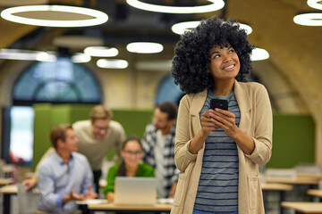 Wall Mural - Young happy mixed race woman holding smartphone, looking aside and smiling while standing in the modern coworking space with her colleagues working on the background